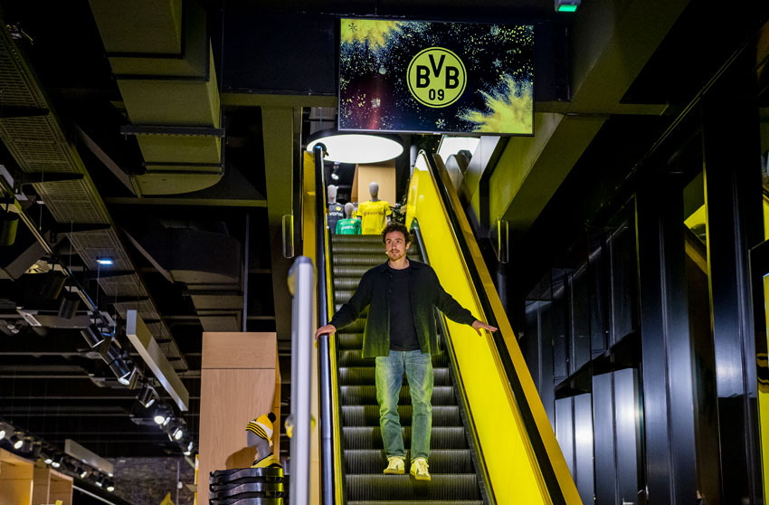 Ein Mann steht auf einer gelben Rolltreppe in einem modernen Gebäude und lächelt in die Kamera.