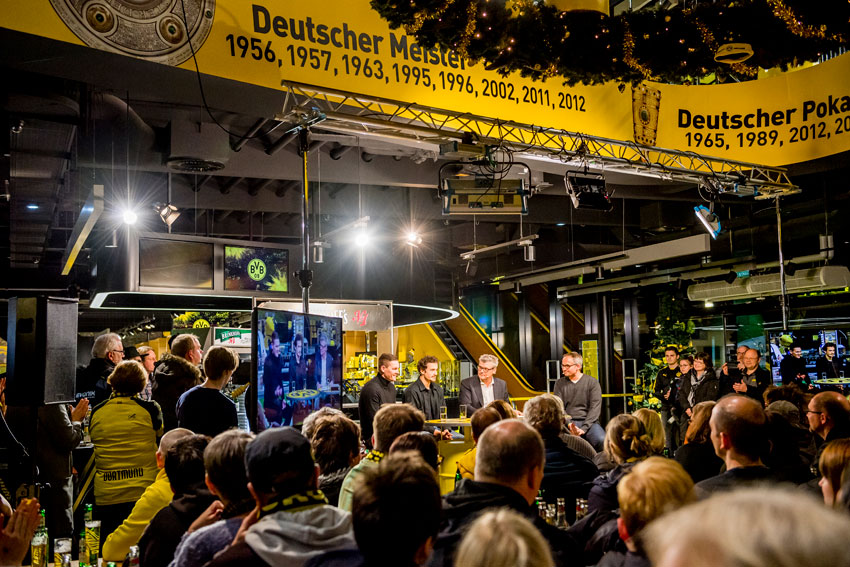 Eine Gruppe Menschen hört bei einem Event in einem beleuchteten Raum vier Personen am Podium zu.
