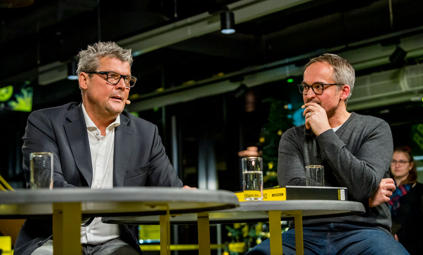 Zwei Männer sitzen an einem Tisch und führen ein Gespräch vor Publikum.
