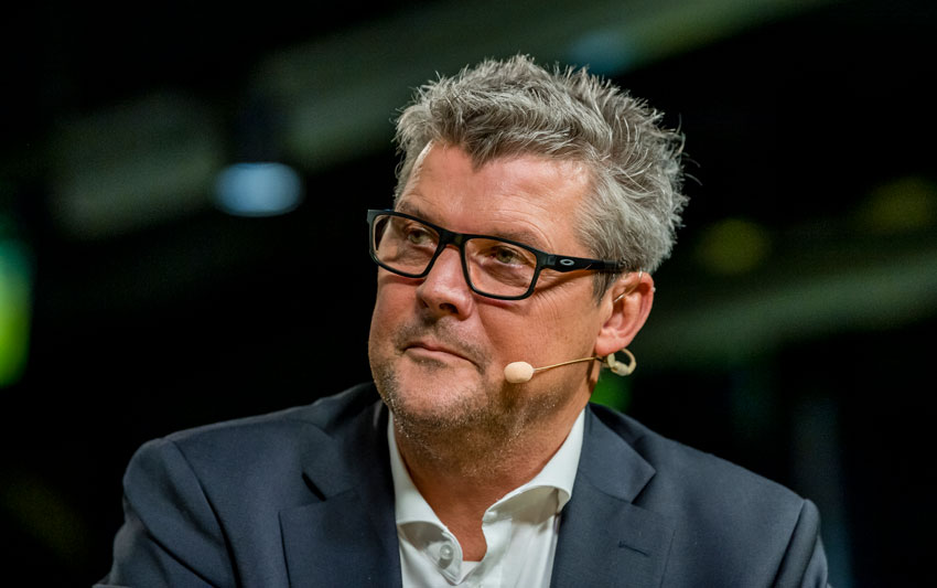 Ein Mann mittleren Alters mit Brille und Mikrofon spricht bei einer Veranstaltung.