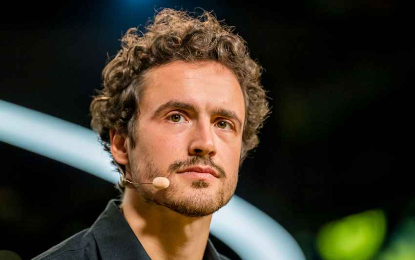 Ein Mann mit lockigem Haar und Mikrofon-Headset spricht bei einer Veranstaltung.