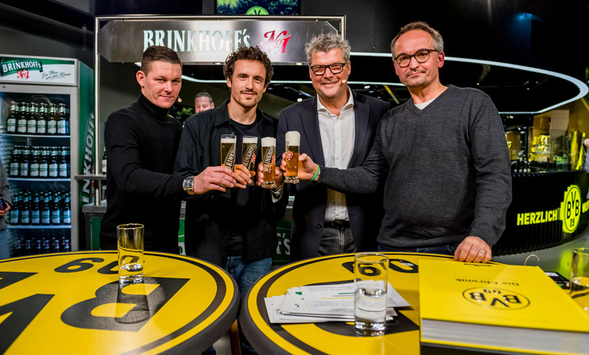 Vier Männer stehen an einem runden Tisch und halten Biergläser hoch.