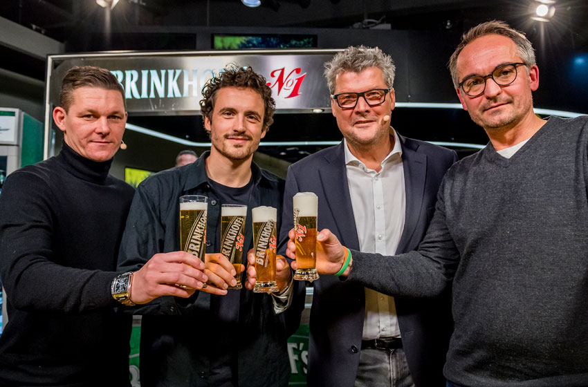 Vier Männer stehen nebeneinander und halten Biergläser in die Kamera. Sie lächeln freundlich.