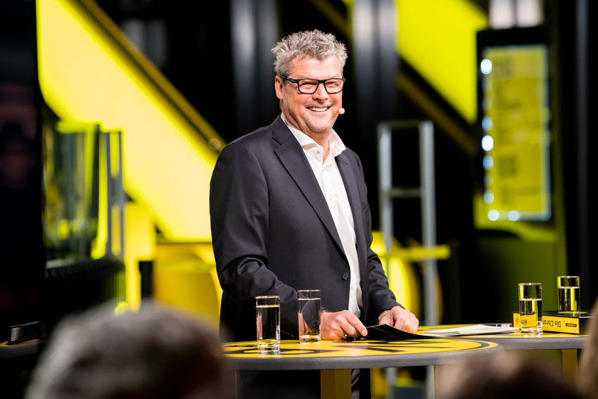 Ein lächelnder Mann in Anzug steht an einem Pult in einem Studio mit gelben Elementen.
