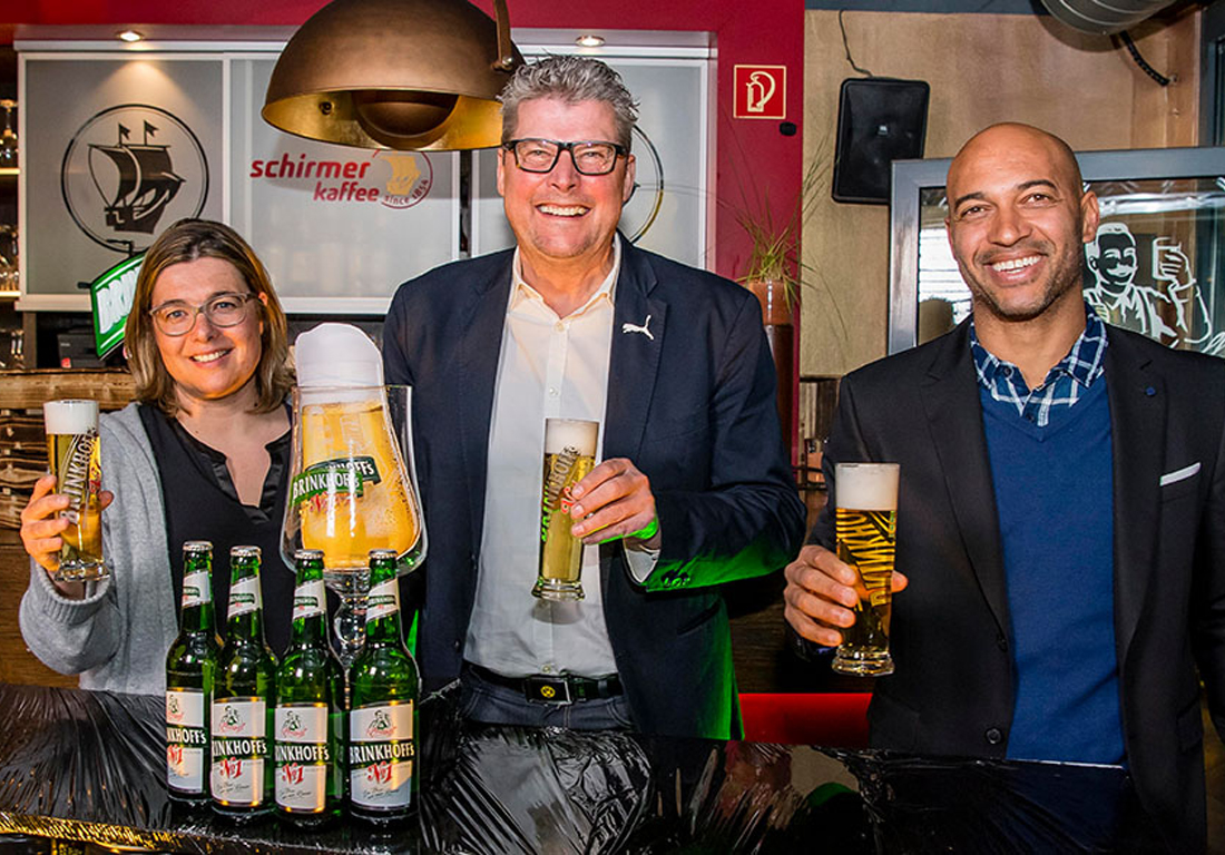 Drei Personen stehen lächelnd in einer Bar und halten Biergläser. Die Frau in der Mitte präsentiert eine große Bierglasattrappe.