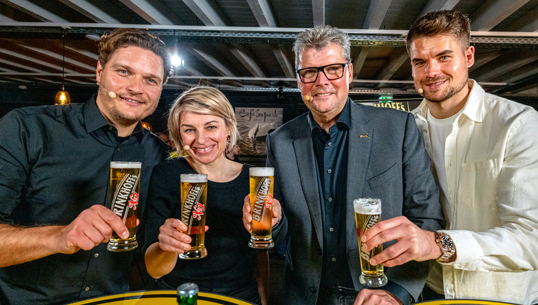 Zwei Männer und zwei Frauen lächeln und halten Biergläser in einer Bar. Sie tragen stilvolle Kleidung.