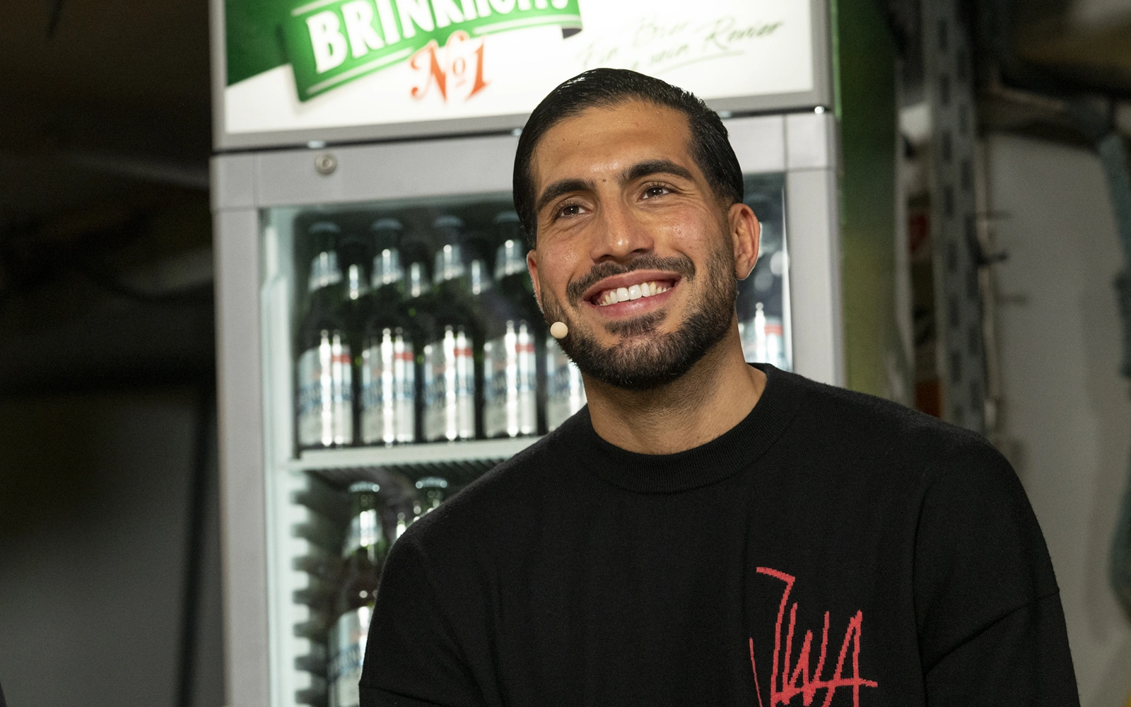 Ein lächelnder Mann mit Bart steht in einem Raum vor einem Getränkekühlschrank.