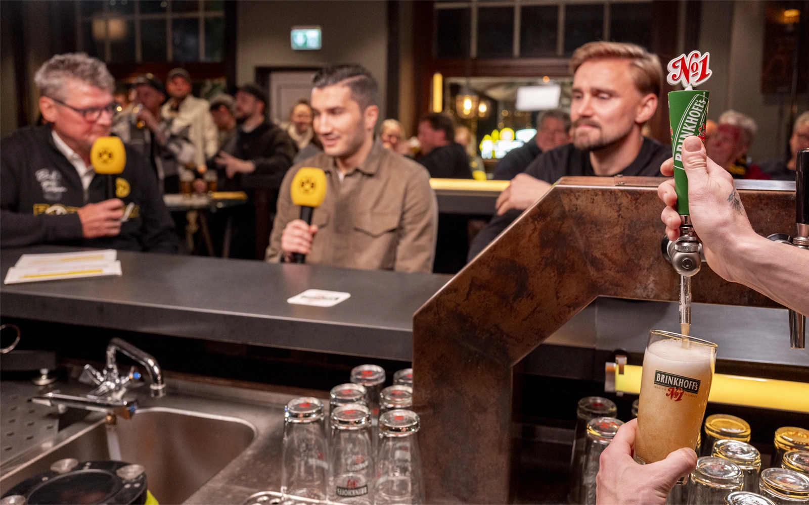 Eine Person zapft Bier in ein Glas, während zwei Männer mit Mikrofonen sprechen.