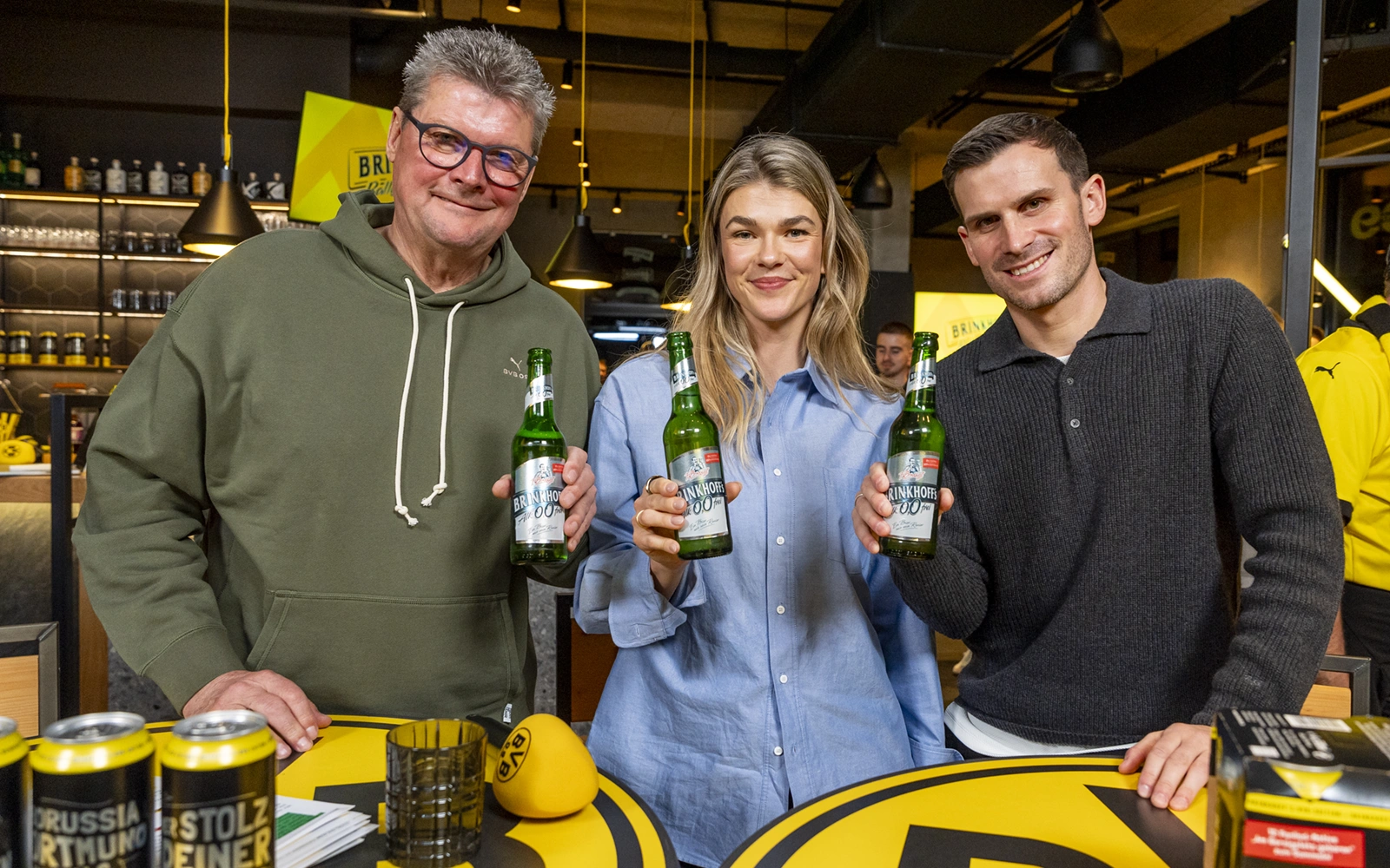 Drei Personen stehen lächelnd in einer Bar und halten je eine Flasche Bier.