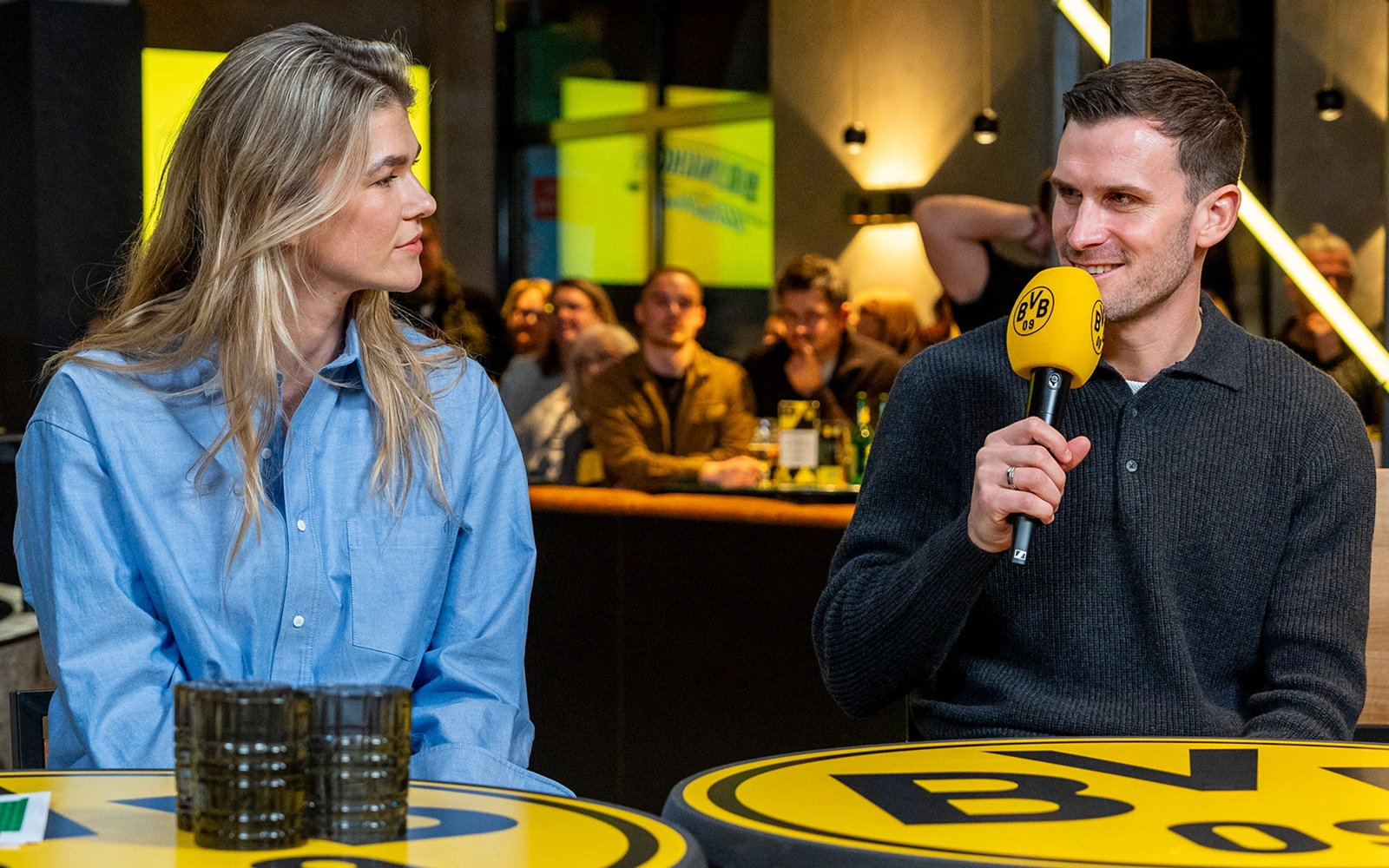 Ein Mann und eine Frau sitzen an einem Tisch und führen ein Interview. Der Mann hält ein Mikrofon.