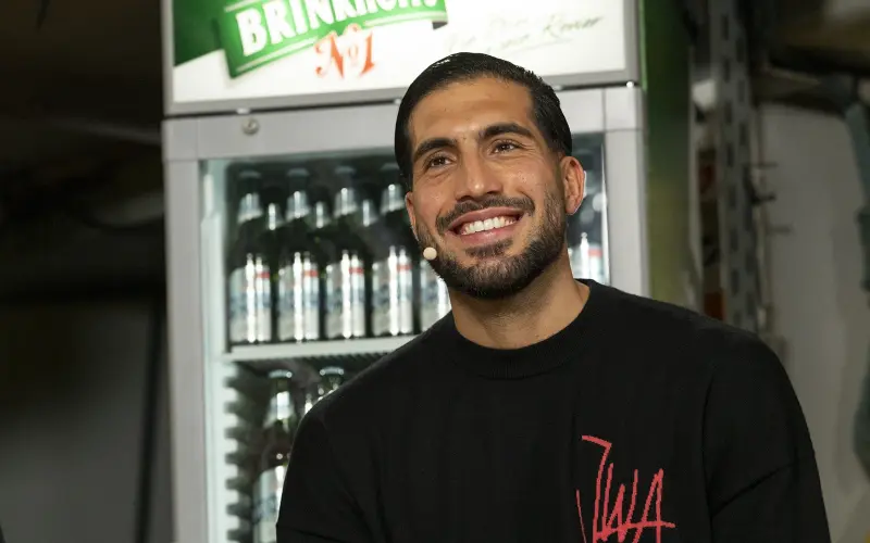 Ein lächelnder Mann mit Bart steht in einem Raum vor einem Getränkekühlschrank.