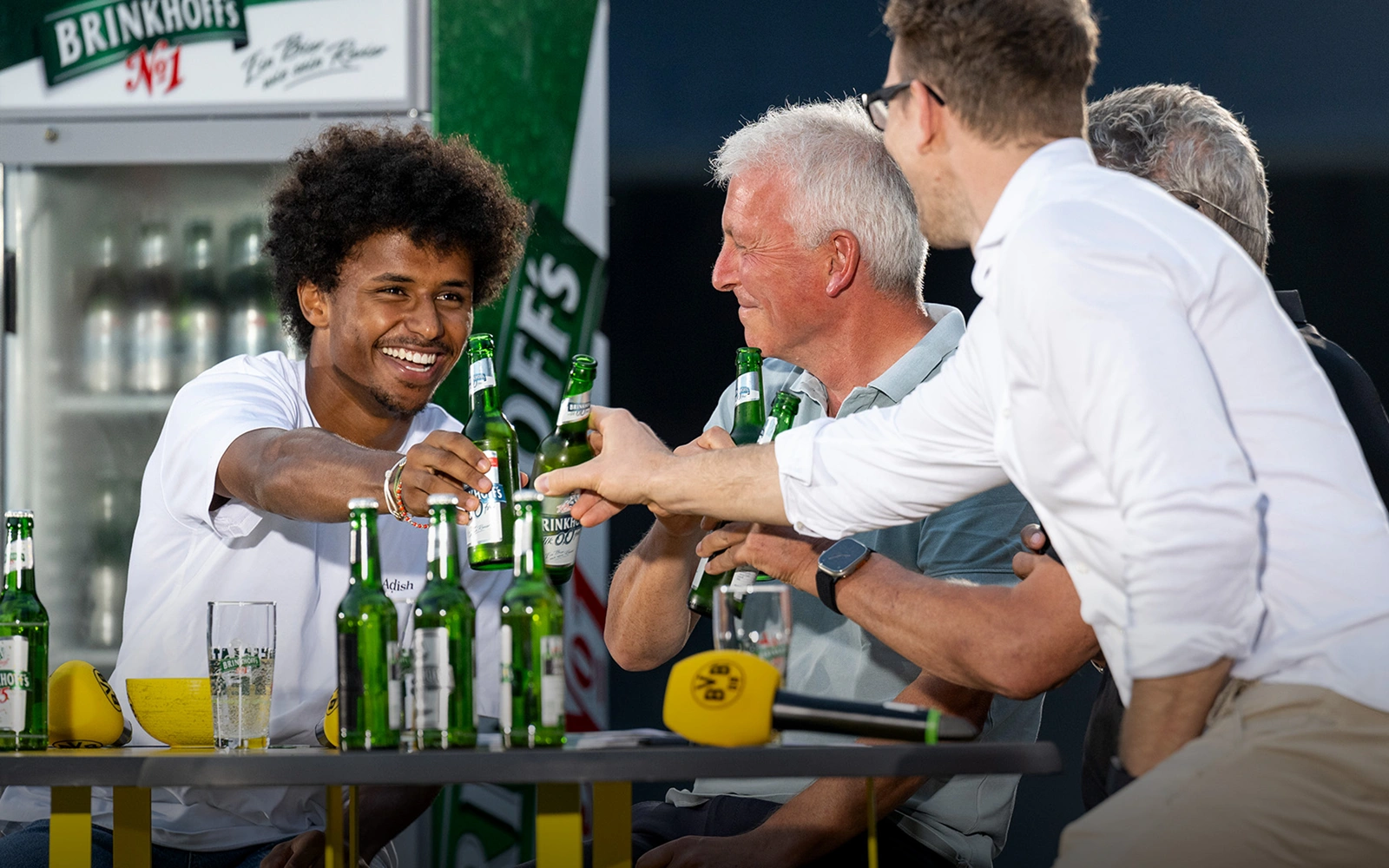 Vier Personen stoßen fröhlich mit grünen Flaschen an einem Tisch an.