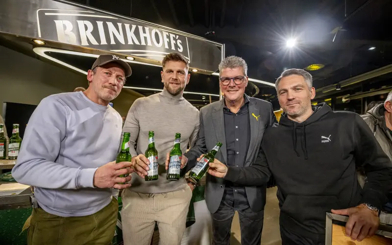 Vier Männer stehen lächelnd an einer Bar und halten Flaschen in den Händen. Sie tragen lässige Kleidung.