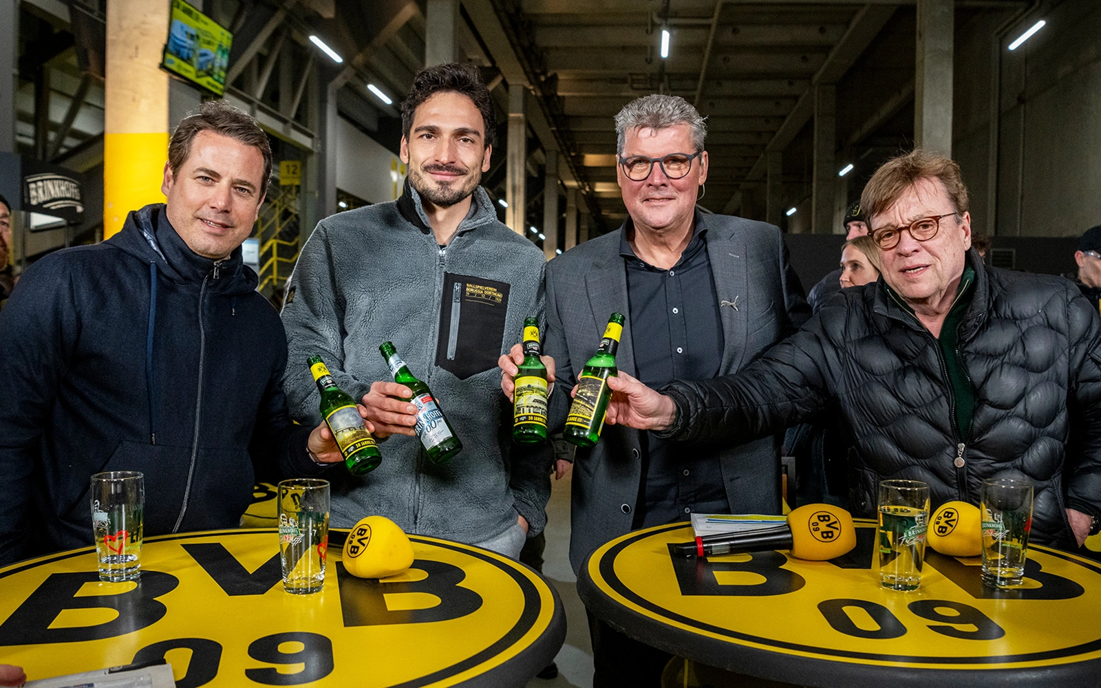 Vier Männer stehen um einen gelben Tisch und halten grüne Flaschen. Sie lächeln in die Kamera.