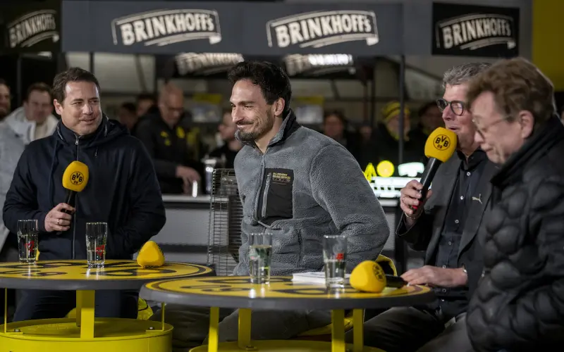 Vier Männer sitzen während einer Radiosendung an einem gelben Tisch und unterhalten sich.