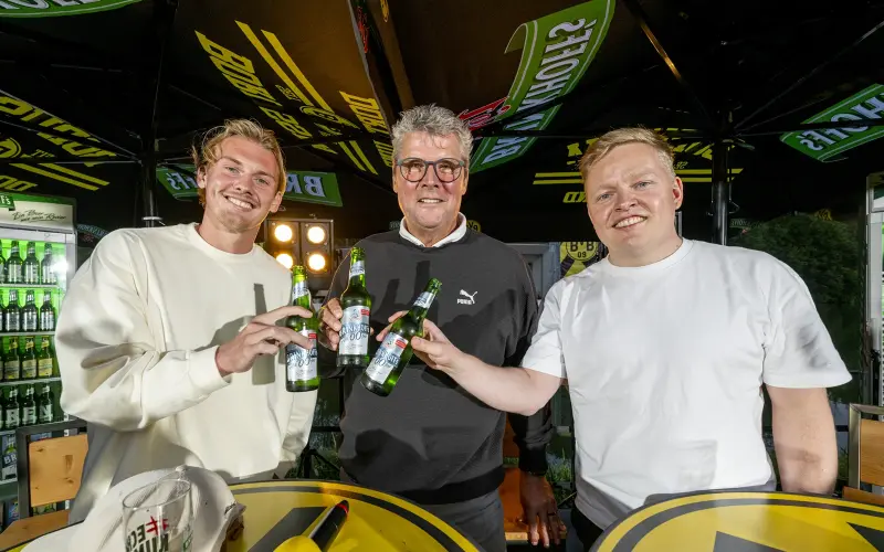 Drei Männer stehen lächelnd an einem Tisch und halten Getränke in den Händen.