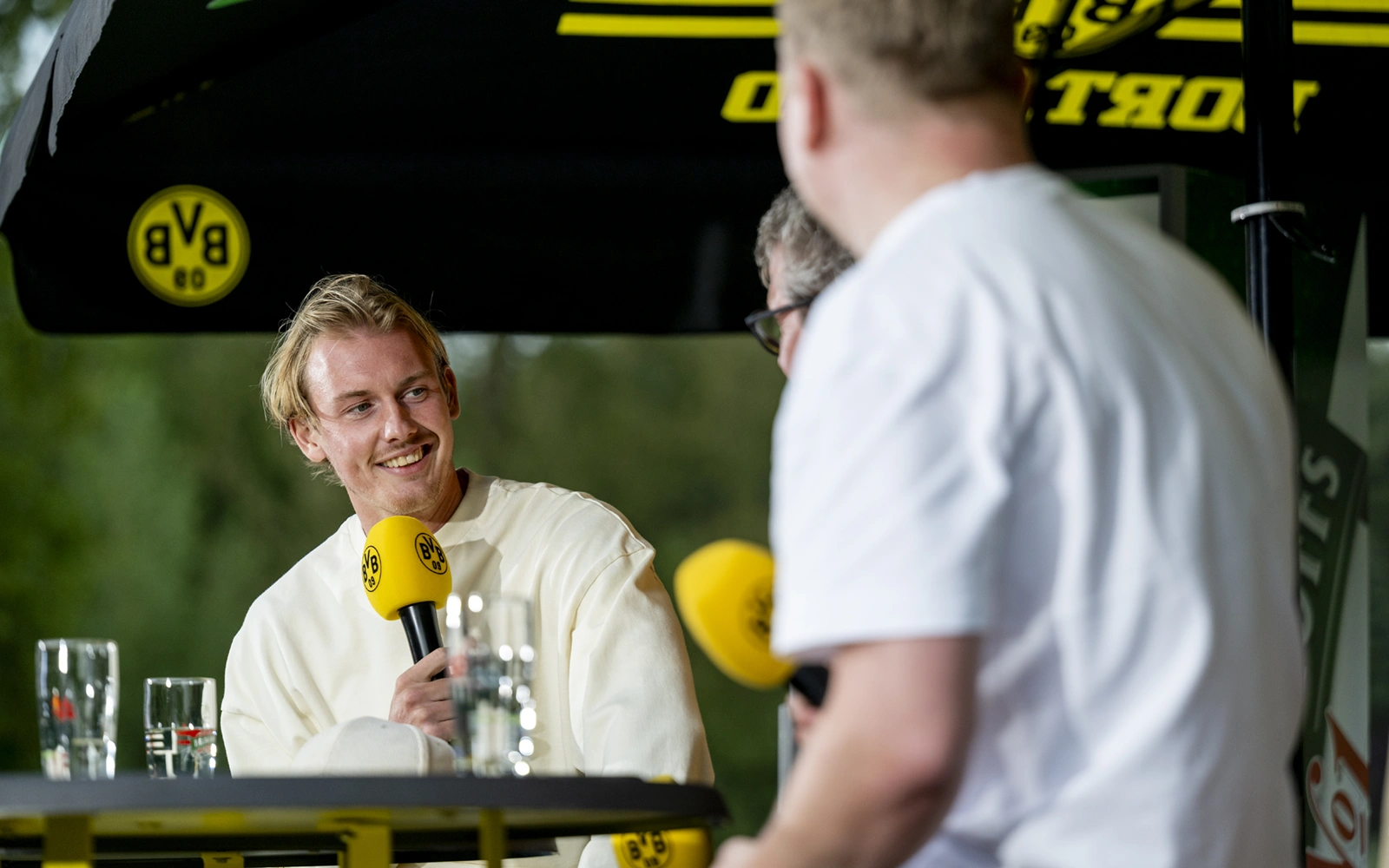 Ein Mann in hellem Oberteil hält ein Mikrofon und spricht lächelnd. Zwei Personen hören ihm zu.