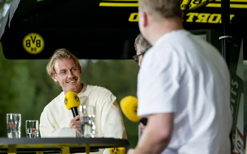 Ein Mann in hellem Oberteil hält ein Mikrofon und spricht lächelnd. Zwei Personen hören ihm zu.