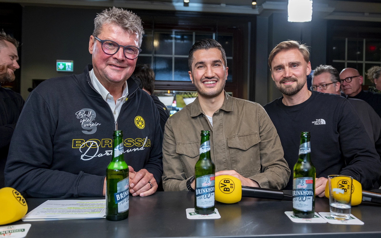 Drei lächelnde Männer sitzen an einem Tisch mit Mikrofonen und Getränken.