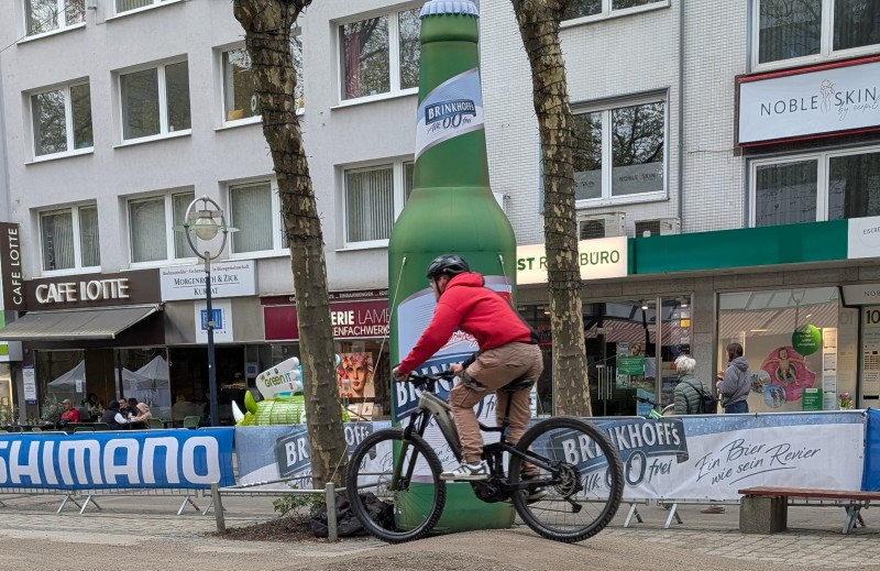 Eine Person fährt Fahrrad auf der Straße, im Hintergrund Häuserfassaden und Bannerwerbung