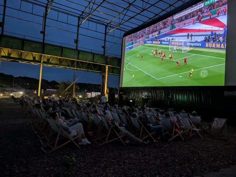 Eine Menschenmenge vor einer großen Leinwand, im Hintergrund der Abendhimmel