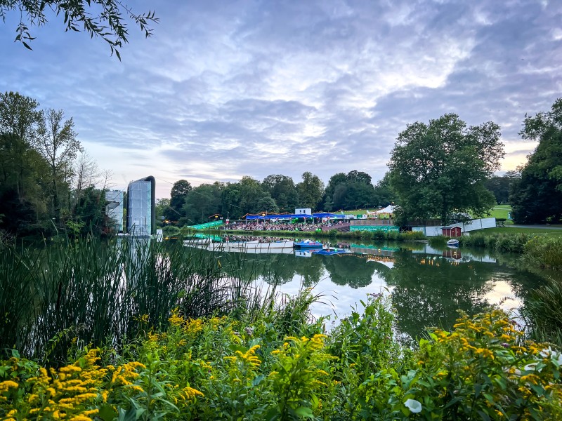Blick auf eine Tribühne in einem viel bewachsenen Park mit Wasserzugang. Auf dem See eine schwimmende Leinwand.