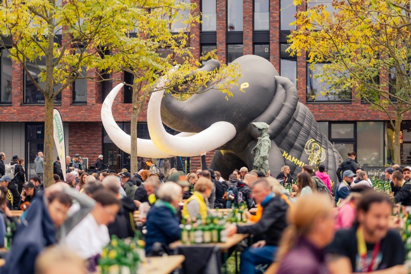 Personen auf Bierbänken, im Hintergrund ein aufblasbares Mammut