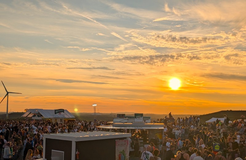 Menschenmenge auf einem Festival mit Ausschankwagen vor einem Sonnenuntergang