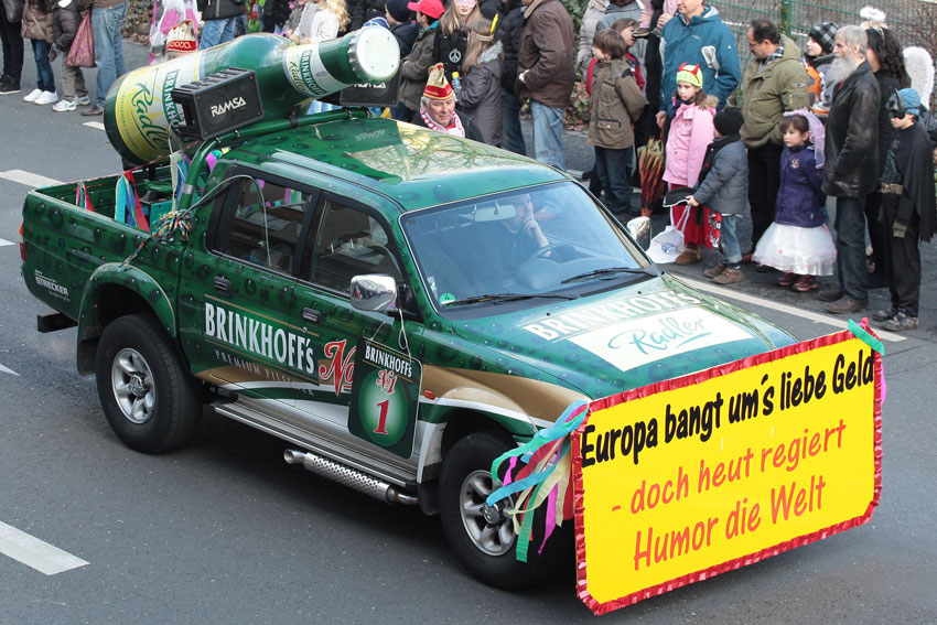Ein grün-brauner Pick-up-Truck ist mit großen Fässern dekoriert und fährt durch eine Menschenmenge.