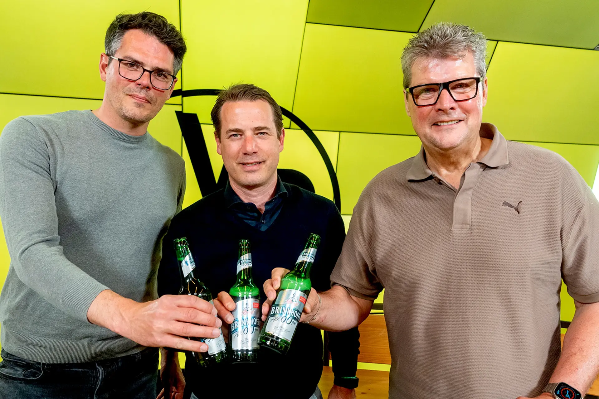 Drei Männer stoßen mit Brinkhoff’s Bierflaschen an und blicken in die Kamera. Im Hintergrund eine Wand mit schwarzem BVB Logo.